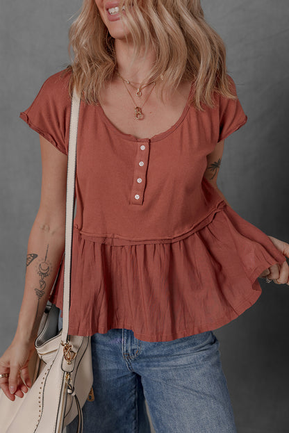 Woman wearing a rust-colored peplum top and blue jeans, holding a beige handbag against a gray background.